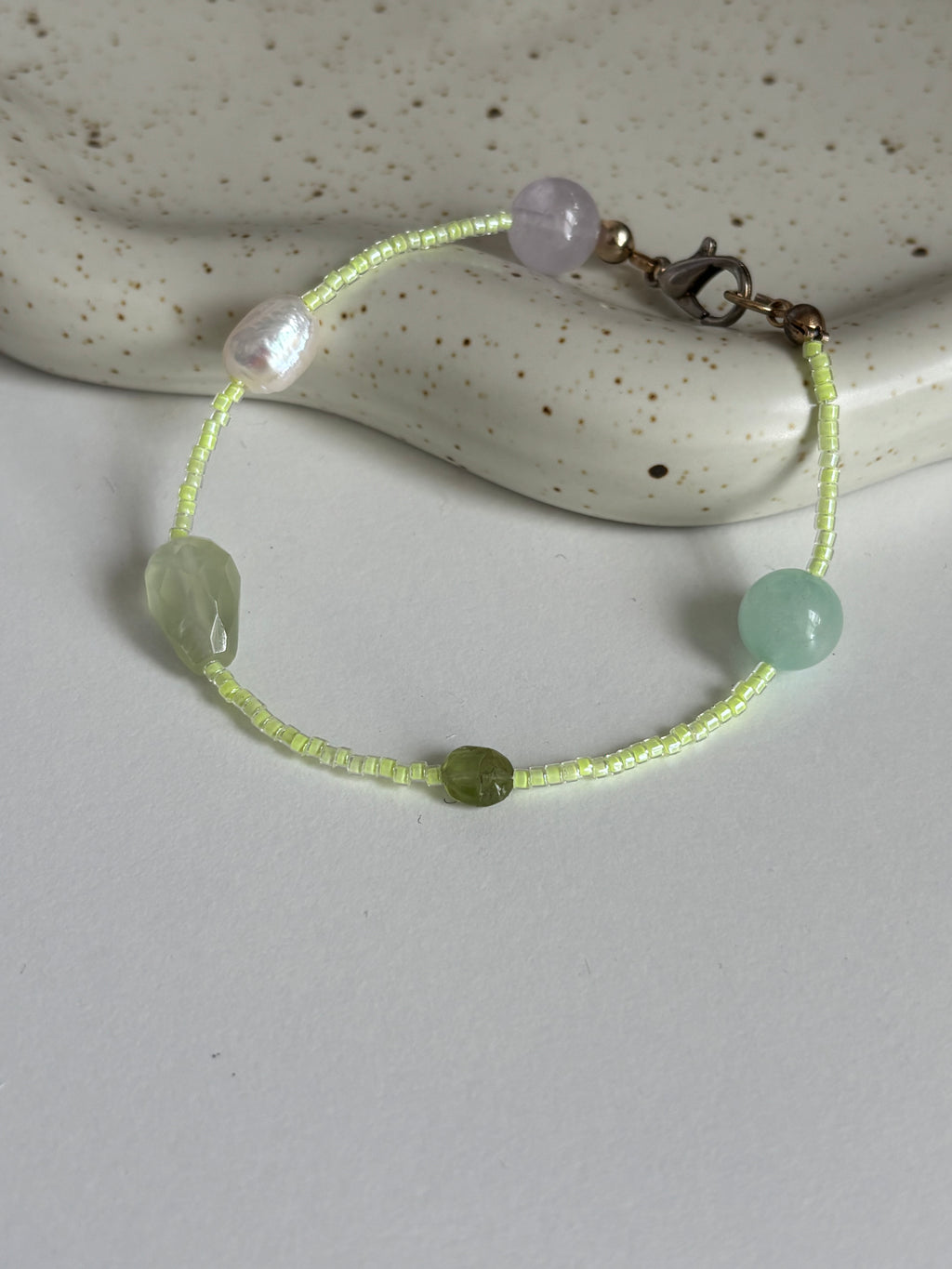 A delicate beaded bracelet with light green and pale lavender beads, resting on a white surface with a speckled ceramic dish in the background.