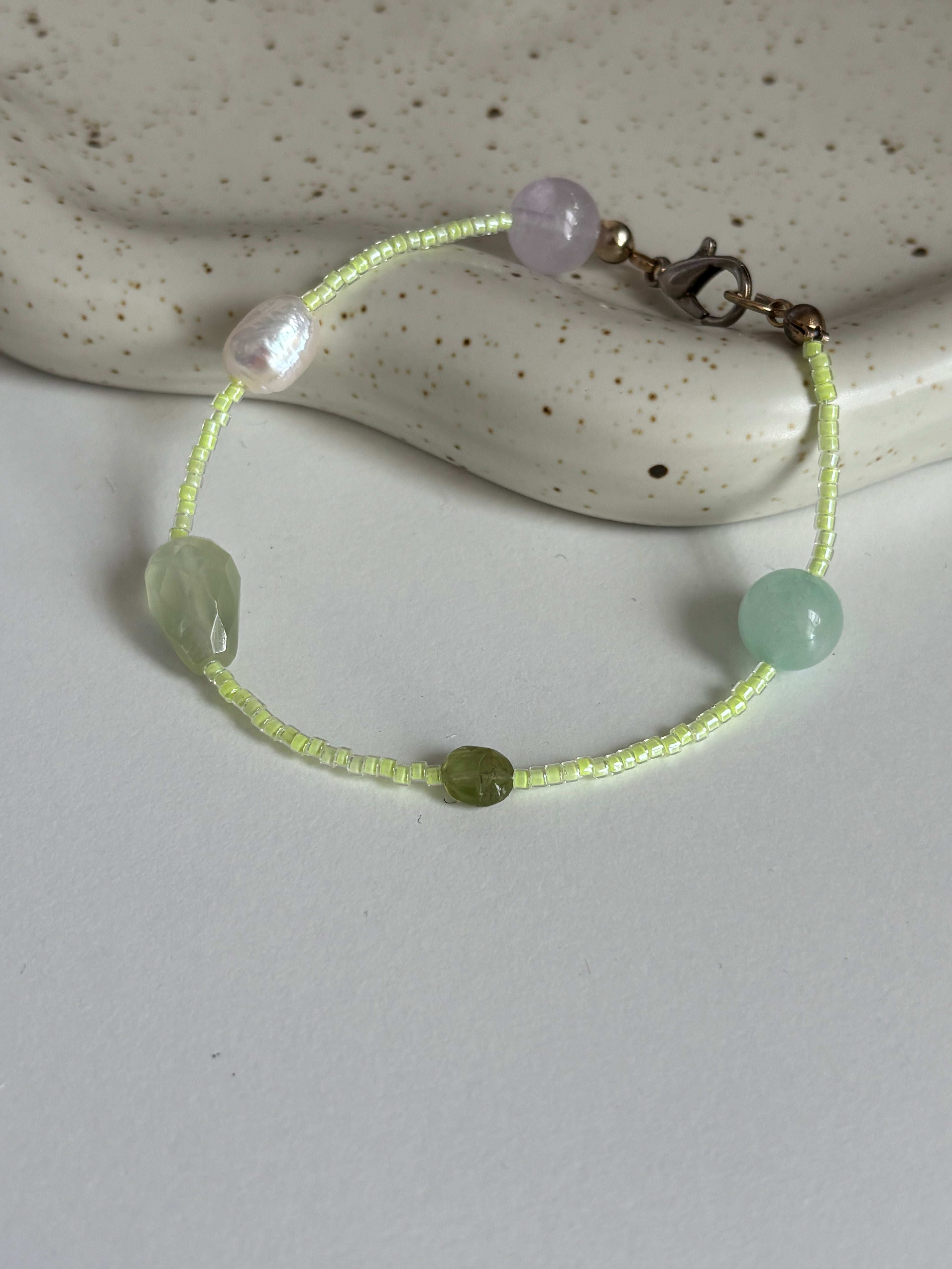 A delicate beaded bracelet with light green and pale lavender beads, resting on a white surface with a speckled ceramic dish in the background.